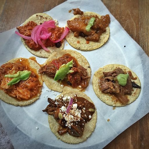 Seis tacos de guisado variados sobre papel: cochinita con cebolla morada, cerdo en salsa, tinga, deshebrada y mole con queso